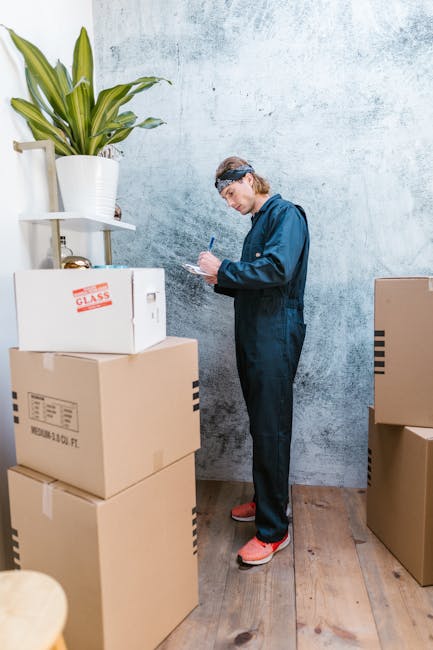 A woman with long blonde hair, wearing a dark grey T-shirt, is standing indoors beside a stack of cardboard packing boxes, some open and others closed, on a wooden floor. She is holding a white clipbo