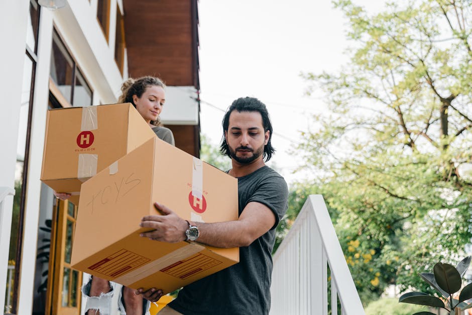 A young woman with curly dark hair, wearing a white T-shirt, blue jeans, and white sneakers, is standing in the doorway of a brick building, carrying a large cardboard box. She has a red and black che